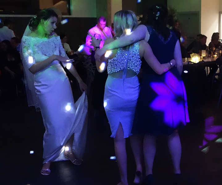 Harriet dances with two of her female wedding guests. This photo shows Harriet on the left, holding up the train of her dress and dancing towards her two friends who are arm in arm with their backs towards us. The woman in the middle is wearing a dress with a sparkly top and the woman on the right has one of R2 Events' flower patterned lights projected onto her dress. The scene is also lit with R2 Event's 'white dots' disco lights.